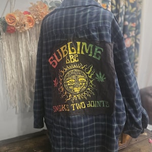 May include: A dark blue and green plaid flannel shirt featuring a large black fabric patch sewn onto the back. The patch displays the text SUBLIME LBC and SMOKE TWO JOINTS in a gradient font, surrounding a yellow sun graphic with leaf icons.