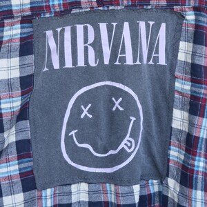 May include: A long-sleeve plaid shirt featuring a dark gray rectangular patch sewn onto the back. The patch displays the word NIRVANA in light purple capital letters above a light purple graphic of a smiley face with X eyes and a tongue out.