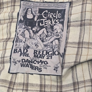 May include: A plaid shirt with a patch featuring the text "Circle Jerks" and "Bad Religion" with concert details. The patch has a black border and a black and white illustration of a nun and other figures.