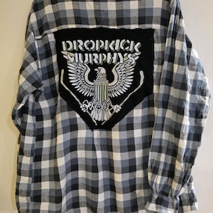May include: A long-sleeved, button-down shirt with a black, white, and gray plaid pattern. The back features a black patch with the words "DROPKICK MURPHYS" above an eagle graphic with crossed hockey sticks.