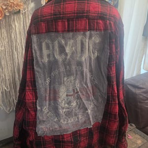 May include: A red and black plaid flannel shirt featuring a large gray rectangular patch sewn onto the back. The patch displays the AC/DC logo, the text "For Those About To Rock We Salute You," and a graphic of a bulldog with a Union Jack flag.