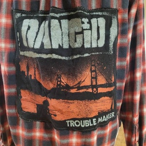 May include: A red and black plaid flannel shirt with a black patch on the back. The patch features a band logo with the word "Rancid" and the text "Trouble Maker".
