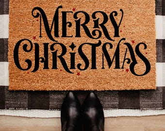 May include: A rectangular doormat with the words "MERRY CHRISTMAS" in large, black, decorative font. Small red stars are scattered around the text. The mat is made of a light brown material and is placed on a black and white checkered rug.