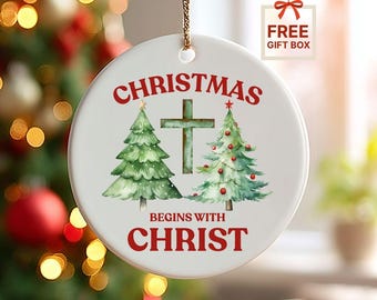 May include: White ceramic Christmas ornament with a gold string. The ornament features a watercolor design of two green Christmas trees, a cross, and the text "CHRISTMAS BEGINS WITH CHRIST" in red.