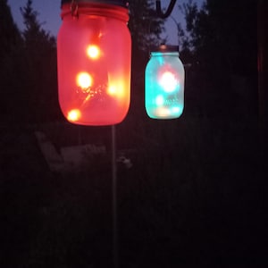 May include: Two mason jar lanterns hanging from a black metal hook. One jar glows red, the other glows teal. Both jars have silver lids and contain small, warm-toned lights. The background is a dark, outdoor setting with trees.