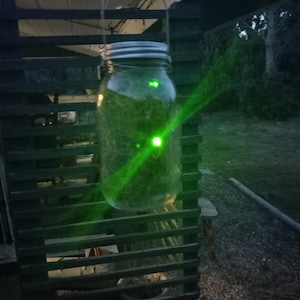 May include: A clear glass mason jar with a metal lid, filled with green material, hangs from a wire. A bright green light shines through the jar, creating a glowing effect. The jar is suspended in front of a wooden structure.