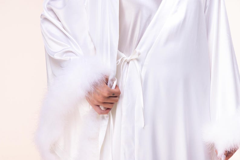 May include: White satin robe with fluffy feather trim on the sleeves. The robe is tied at the waist with a matching satin belt. The sleeves are long and the robe appears to be made of a soft, flowing fabric.
