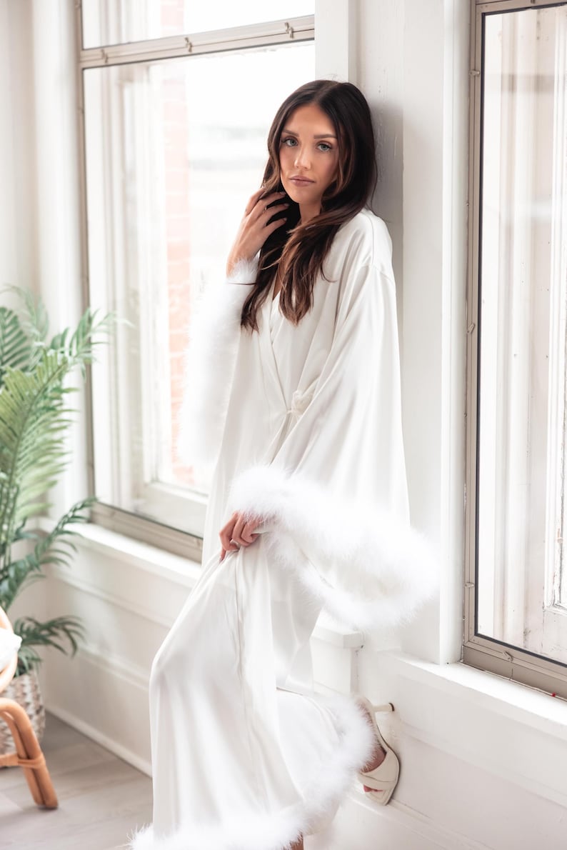 May include: A white satin robe with fluffy feather trim on the sleeves and hem. The robe is open, revealing a glimpse of the wearer's legs and a pair of white heels. The model is leaning against a white window frame.
