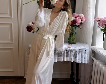May include: A floor-length cream-coloured robe featuring long sleeves with white feather trim at the cuffs and a matching white feather trim along the bottom hem. The robe includes a wrap-style front with a tie belt at the waist and a deep V-neckline.