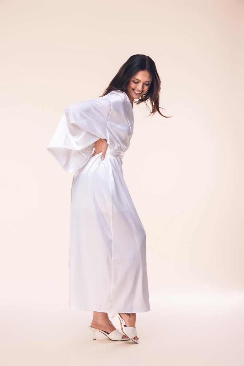 May include: A long, flowing white satin robe with wide sleeves and a matching belt. The robe is worn with white heeled sandals. The model is smiling and has dark hair.