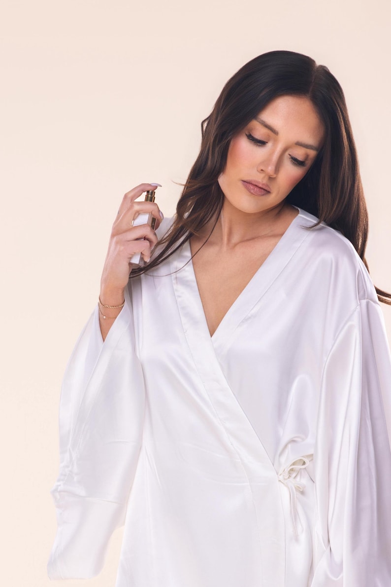 May include: A white satin robe with wide sleeves and a wrap-around design. The robe is tied at the waist with a matching fabric belt. A small perfume bottle is held in the hand.