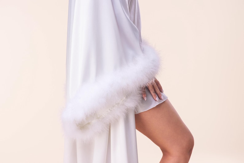 May include: A white satin robe with fluffy white feather trim on the sleeves. The robe is draped, with a glimpse of a matching slip underneath. The fabric has a soft, flowing appearance.