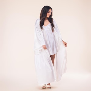 May include: A woman wearing a white satin robe and matching slip dress. The robe has long, flowing sleeves and is open in the front. The slip dress has a V-neck. The woman is also wearing white sandals.
