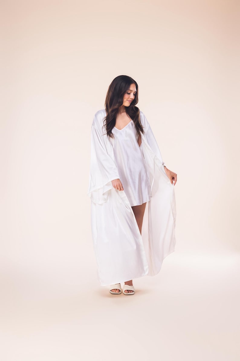 May include: A woman wearing a white satin robe and matching slip dress. The robe has long, flowing sleeves and is open in the front. The slip dress has a V-neck. The woman is also wearing white sandals.