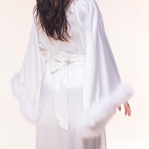 May include: White satin robe with wide sleeves and a matching tie belt. The robe features fluffy white feather trim on the sleeve cuffs. The back of the robe has a bow detail at the waist.
