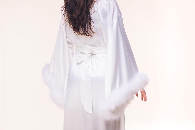 May include: White satin robe with wide sleeves and a matching tie belt. The robe features fluffy white feather trim on the sleeve cuffs. The back of the robe has a bow detail at the waist.