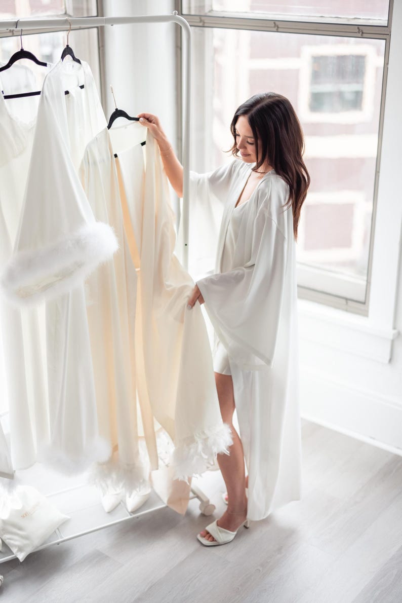 May include: A selection of white robes and loungewear hanging on a clothing rack. One robe has a cream color and feather trim. The other robes are white. The scene is in a bright room with a window.