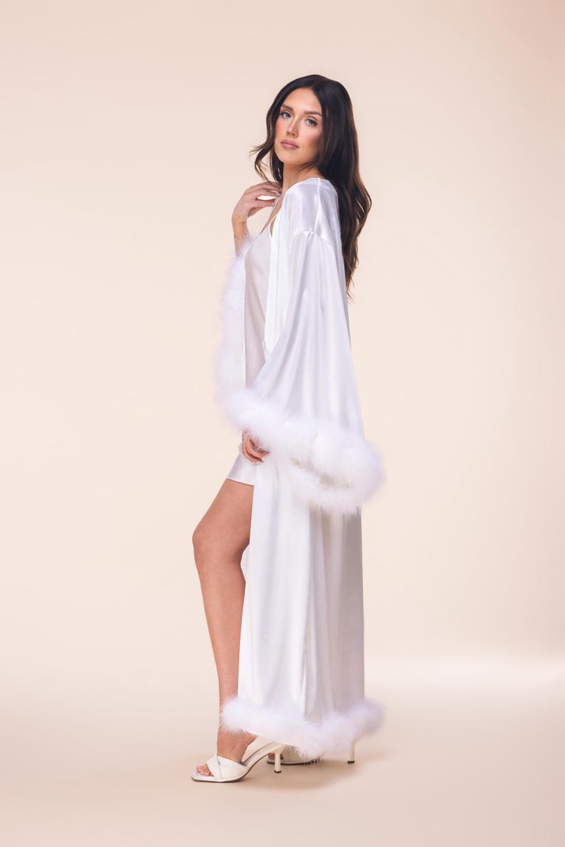 May include: A long, white satin robe with fluffy white feather trim on the sleeves and hem. The robe is worn over a matching white slip dress. The model is wearing white high-heeled sandals.