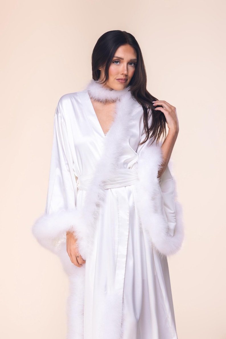 May include: A white satin robe with feather trim on the cuffs and lapels. The robe has a matching feather collar and a tie belt. The long-sleeved robe is a luxurious garment.