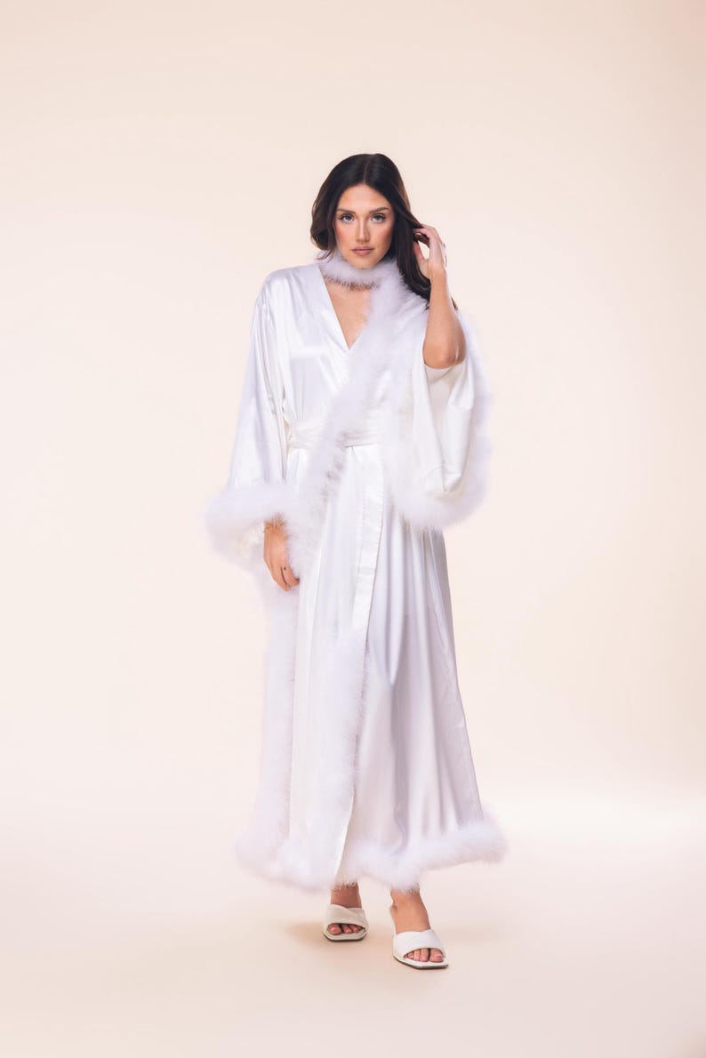 May include: A white satin robe with feather trim on the cuffs, hem, and collar. The robe is long-sleeved and has a matching belt. The model is wearing white sandals. The robe is a luxurious item, perfect for lounging or bridal wear.