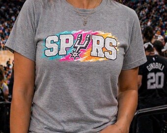 May include: A gray t-shirt with the word "SPURS" in white block letters with a colorful paint stroke design. The shirt features the Spurs logo and is worn with blue jeans. A person is wearing the shirt.