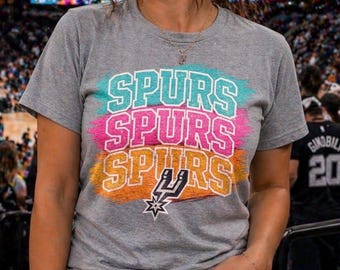 May include: A gray t-shirt with the word "SPURS" stacked three times in teal, pink, and orange, with a team logo at the bottom. The shirt is short-sleeved and worn by a person in a stadium setting.