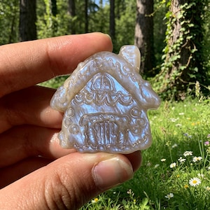 Könnte beinhalten: Eine geschnitzte Steinfigur in Form eines kleinen Hauses mit Schornstein, die eine glatte, schillernde Oberfläche aufweist, welche Licht in Blau- und Silbertönen reflektiert. Das Haus hat geätzte Details wie eine zentrale Tür und Fenster.