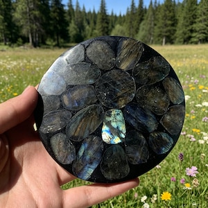 May include: A circular black coaster or decorative disc inlaid with multiple polished labradorite stones. The stones feature dark grey and black tones with iridescent flashes of blue, gold, and green light reflecting from their surfaces.