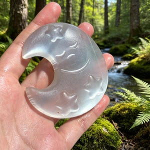 May include: A translucent, crescent moon-shaped object carved from white selenite crystal. The surface features an engraved, smiling face with closed eyes and three embossed five-pointed stars arranged around the moon's curved body.