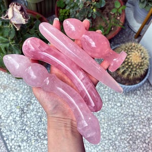 Peut inclure: Une collection d'outils de massage en quartz rose, de formes variées, dont des baguettes et des formes bulbeuses. Les pierres polies présentent différentes tailles et formes, avec des nuances de rose. Les outils sont tenus dans une main, avec des plantes en arrière-plan.
