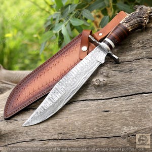 May include: A Damascus steel hunting knife with a brown leather sheath. The knife features a patterned blade, a handle with a deer antler design, and a leather strap. The sheath is embossed with a decorative pattern.