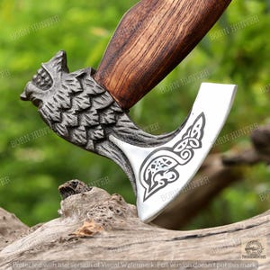 May include: A detailed close-up of a handcrafted axe. The axe head is silver with a Celtic knot design, and the handle is dark wood. The axe features a decorative wolf head carving where the head meets the handle. The text "BORN HUNTER" is visible.
