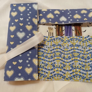 Könnte beinhalten: Eine blaue Stoff-Stricknadel-Tasche mit Herz- und Sternmuster. Die Tasche ist geöffnet und zeigt verschiedene Stricknadeln. Ein weißes Band ist um die Tasche gebunden. Die Innenseite hat ein geometrisches Muster in Blau, Gelb und Weiß.