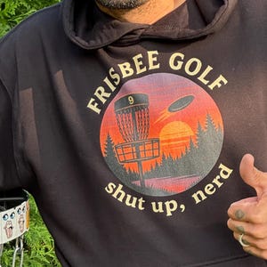 May include: A black hoodie with a "Frisbee Golf" graphic featuring a sunset and disc golf basket. The text "shut up, nerd" is below the graphic. A beige disc and a disc golf basket are also visible.