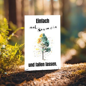 Puede incluir: Una tarjeta rectangular blanca con el texto alemán "Einfach mal Baum sein" y "und fallen lassen". La tarjeta presenta una ilustración de acuarela de un árbol. El fondo es una escena borrosa del bosque con follaje verde.