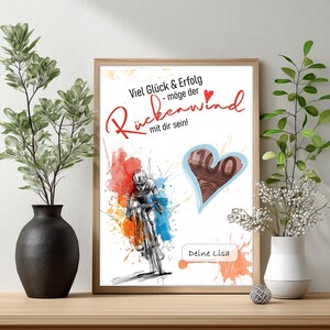 Op de afbeelding: Een ingelijste print met een illustratie van een fietser, levendige verfspatten en Duitse tekst. Het kunstwerk bevat een hartvormig ontwerp. De lijst is warm bruin van kleur en de print wordt getoond op een houten oppervlak.