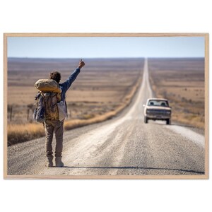 Op de afbeelding: Een reiziger met een grote beige rugzak staat op een grindweg en steekt een arm uit om te liften. Een lichtgekleurde pick-up nadert in de verte op de lange, rechte weg die door een uitgestrekt, dor en open landschap loopt.