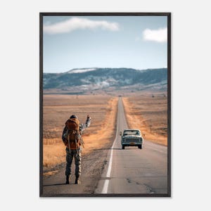 Op de afbeelding: Een ingelijste kunstdruk met een reiziger met een grote bruine rugzak die op de berm van een lange, rechte asfaltweg staat en liftend met de duim omhoog staat terwijl een vintage lichtgekleurde pick-up in de verte nadert.