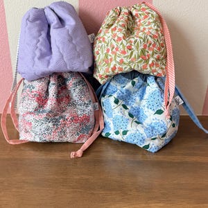 May include: Four drawstring bags in various floral and patterned fabrics. The bags are in shades of lavender, pink, green, and blue. Each bag has a coordinating drawstring closure. The bags are stacked on a wooden surface.