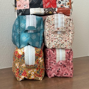 May include: A stack of five small, square fabric pouches. The top pouch is a patchwork of floral and geometric patterns. The other pouches are teal, cream, yellow, and pink, each with a white zipper and a quilted design.
