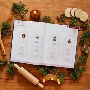 May include: An open book with a blue cover displays a December weekly overview with illustrations of a wreath, house, basket, and Christmas tree. A wooden rolling pin, candle holder, and wooden ornaments are on a wooden surface.