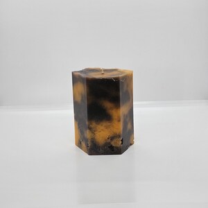 May include: A hexagonal pillar candle with a marbled design in shades of brown and amber. The candle has a rough, textured surface and a single wick at the top. The candle is a decorative item for home decor.