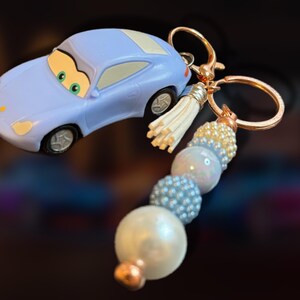 May include: A light blue car-shaped keychain with a gold-colored keyring and a beaded tassel. The keychain features a cartoon car design with green eyes and a white roof. The beaded tassel includes white, pearl, and blue beads.