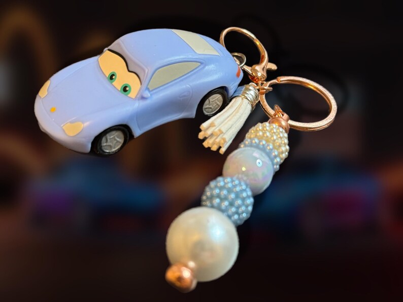 May include: A light blue car-shaped keychain with a gold-colored keyring and a beaded tassel. The keychain features a cartoon car design with green eyes and a white roof. The beaded tassel includes white, pearl, and blue beads.