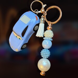 May include: A light blue car-shaped keychain with a gold-colored keyring and beaded accents. The keychain features a small white tassel and a series of pearl and blue beads. The car has black wheels and a white window.