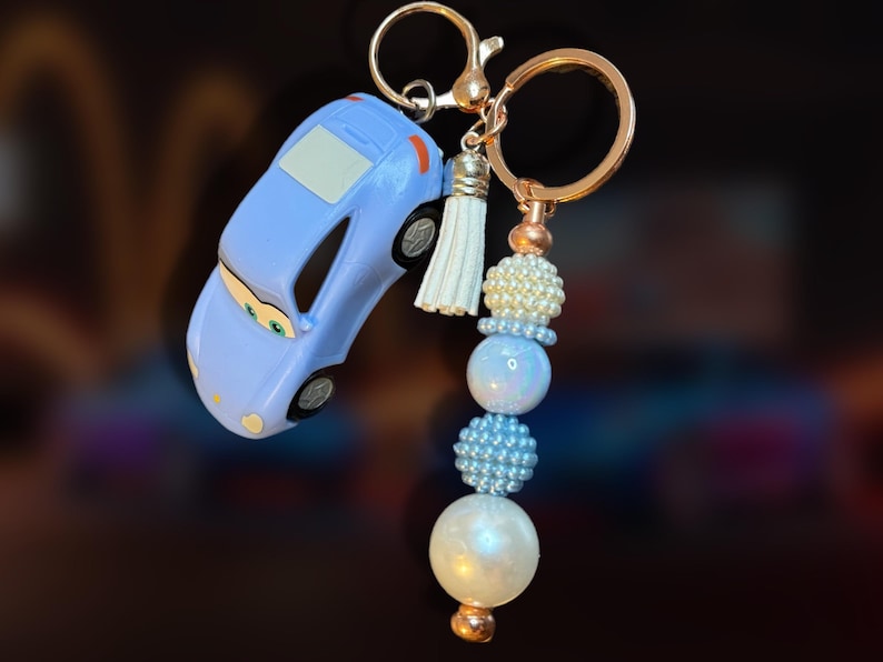 May include: A light blue car-shaped keychain with a gold-colored keyring and beaded accents. The keychain features a small white tassel and a series of pearl and blue beads. The car has black wheels and a white window.