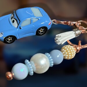 May include: A light blue toy car keychain with a rose gold clasp and a white tassel. The keychain also features a beaded charm with white, light blue, and gold beads. The car has black wheels.