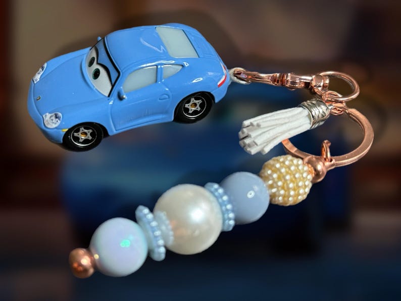 May include: A light blue toy car keychain with a rose gold clasp and a white tassel. The keychain also features a beaded charm with white, light blue, and gold beads. The car has black wheels.