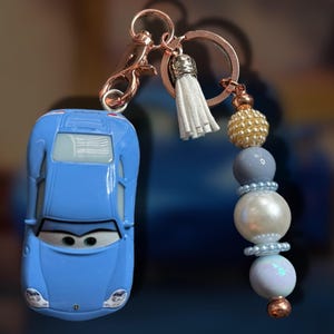 May include: A blue car-shaped keychain with a rose gold clasp and a beaded charm. The charm features a white tassel, gold and blue beads, and a large white pearl-like bead. The keychain is a fun accessory.
