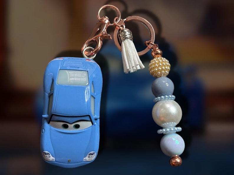 May include: A blue car-shaped keychain with a rose gold clasp and a beaded charm. The charm features a white tassel, gold and blue beads, and a large white pearl-like bead. The keychain is a fun accessory.